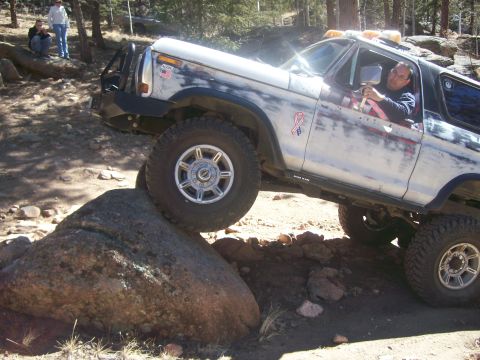 1993 Ford Bronco - A white and blue Ford Bronco is navigating a rocky trail, with its front tire on top of a large boulder. The driver is visible through the…
