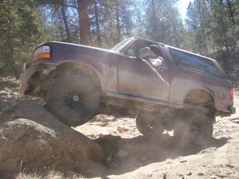 1993 Ford Bronco - A brown 1993 Ford Bronco is shown off-roading on a rocky dirt trail, with the front tire climbing over a large boulder. Dust is kicked up a…