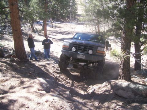 1993 Ford Bronco - A dark-colored Ford Bronco is navigating a rocky, off-road trail in a wooded area. Two men are standing near a tree in the background, obse…