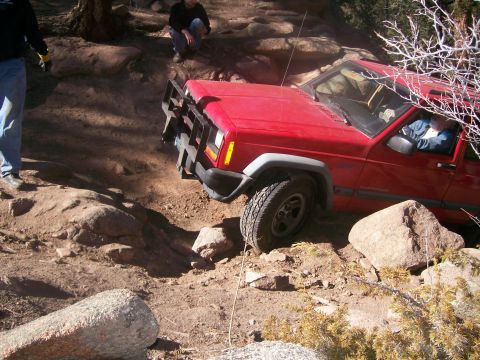 1993 Ford Bronco - A red Jeep Cherokee is navigating a rocky, off-road trail with a custom brush guard. Two people are visible near the vehicle, one standing…