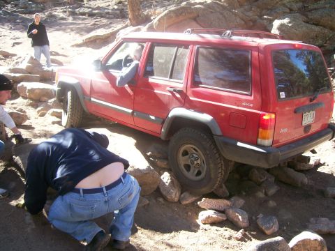 1993 Ford Bronco - A red Jeep Cherokee is navigating a rocky trail, with its rear tires elevated on rocks. Several people are visible around the vehicle, with…