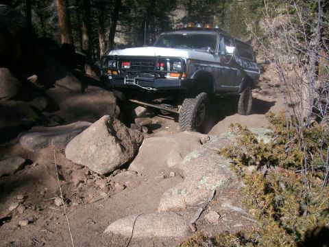 1993 Ford Bronco - A lifted, older model Ford Bronco is navigating a rocky, off-road trail in a wooded area. The vehicle is positioned at an angle, with its f…