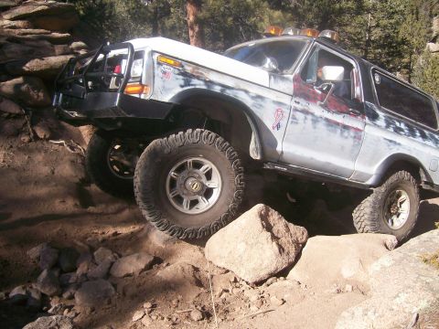 1993 Ford Bronco - A heavily modified, light-colored Ford Bronco is navigating a rocky, off-road trail. The vehicle features aggressive tires, a custom front…