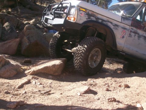 1993 Ford Bronco - A lifted, off-road modified Ford Bronco is shown navigating a rocky terrain. The vehicle features a prominent aftermarket bumper and large,…