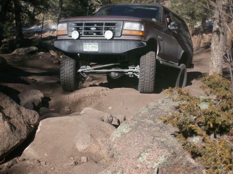 1993 Ford Bronco - A brown 1993 Ford Bronco is shown driving off-road on a rocky trail. The front of the vehicle is angled towards the camera, with its tires…