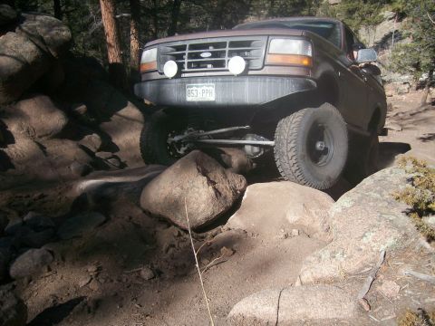 1993 Ford Bronco - A brown 1993 Ford Bronco is navigating a rocky, off-road trail, with its front tire positioned on a large boulder.