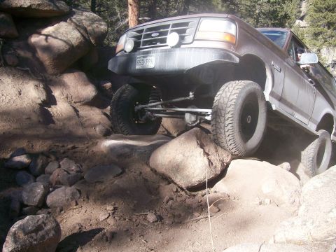 1993 Ford Bronco - A brown Ford Bronco is navigating a rocky, off-road trail. The front tire is lifted high over a large boulder, showcasing the vehicle's sus…