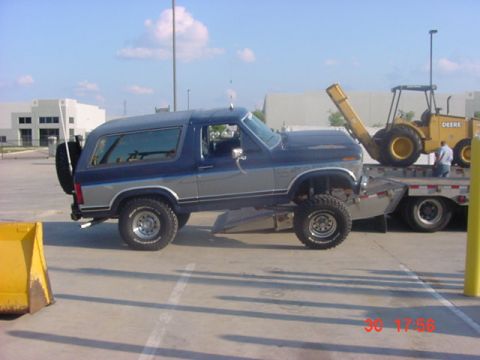 1986 Ford Bronco - A blue and silver Ford Bronco is loaded onto a trailer, with a yellow loader visible in the background.
