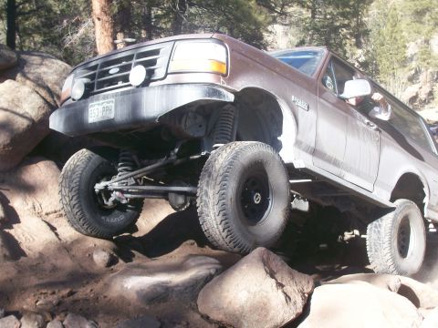 1993 Ford Bronco - A brown 1993 Ford Bronco is navigating a rocky, off-road trail. The vehicle is angled uphill, with its front left tire significantly elevat…