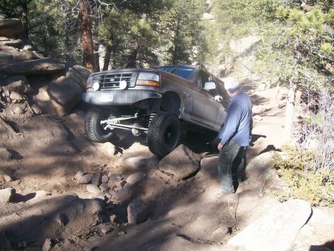 1993 Ford Bronco - A brown 1993 Ford Bronco is navigating a rocky, off-road trail with its front wheels on a steep incline. A man wearing a blue plaid shirt a…