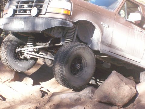1993 Ford Bronco - The front driver's side of a brown Ford Bronco is shown at an upward angle, navigating over large rocks on a dirt trail. The vehicle's susp…