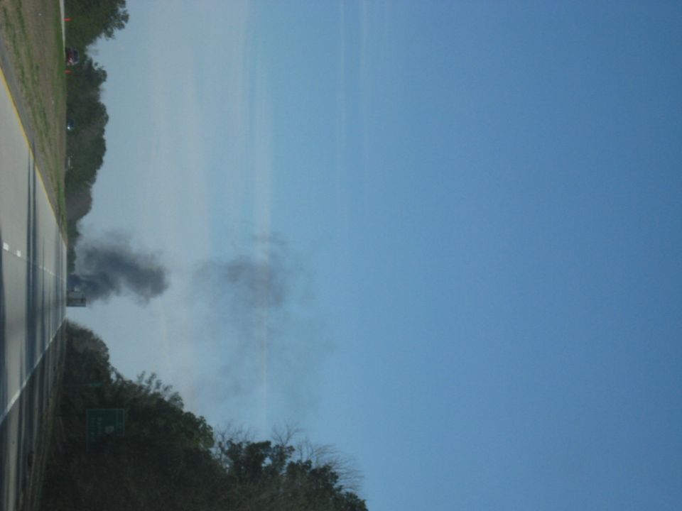 Sideways photo of dark smoke rising into a blue sky beyond roadside trees