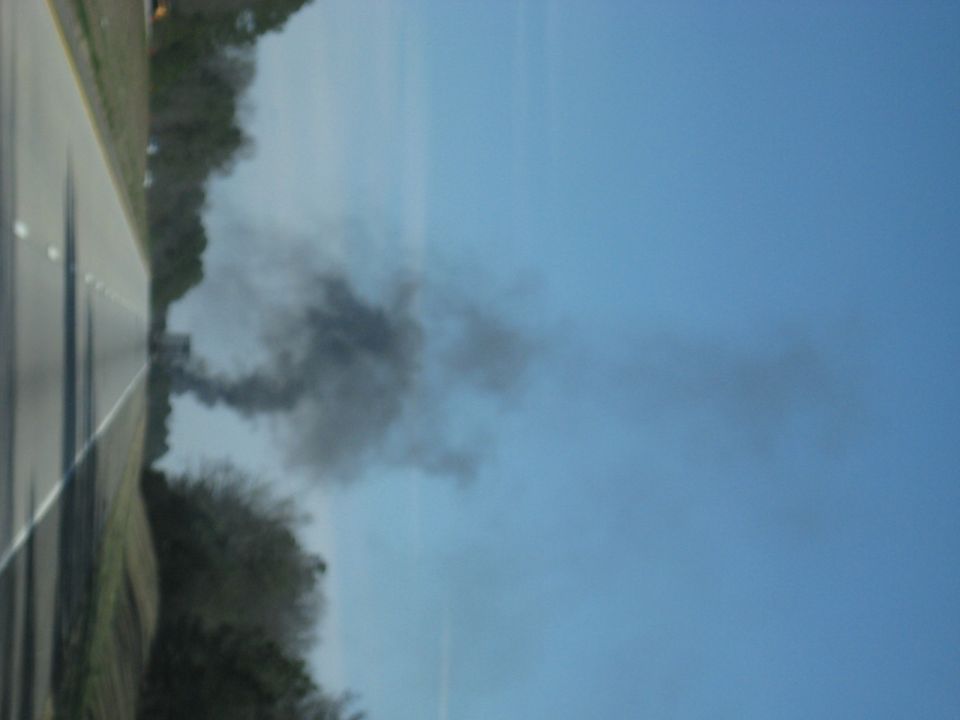 Sideways photo of black smoke drifting over a roadside treeline