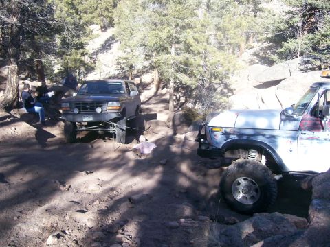 1993 Ford Bronco - Two off-road vehicles are parked on a dirt trail surrounded by trees and rocks. The vehicle on the left is a dark-colored Ford Bronco, and…
