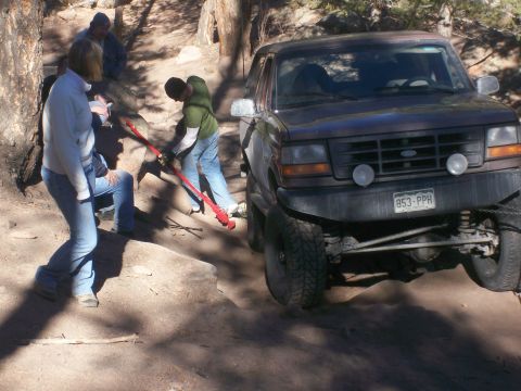 1993 Ford Bronco - A group of people are working on a dark-colored Ford Bronco that appears to be stuck on a dirt trail. One person is using a red jack to lif…