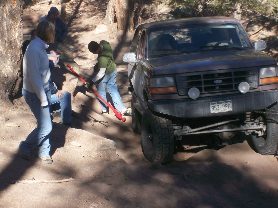 A dark-colored Ford Bronco is being lifted with a red jack on a dirt trail.