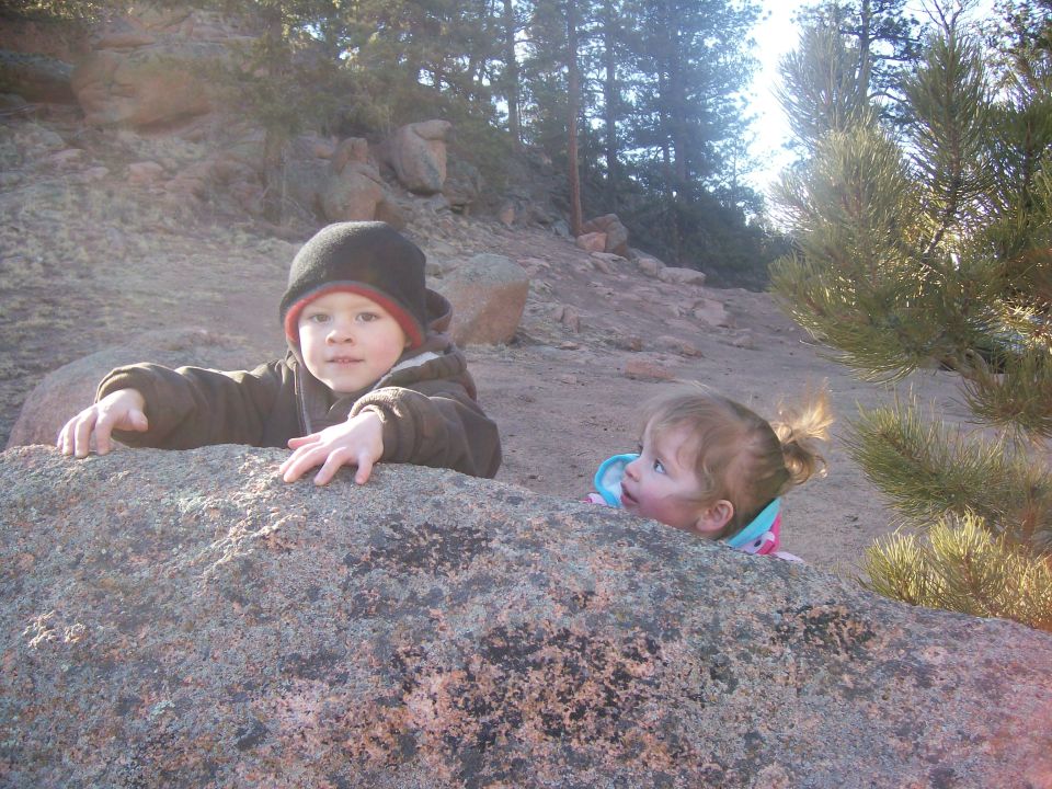 Two young children peeking over a large rock outdoors.