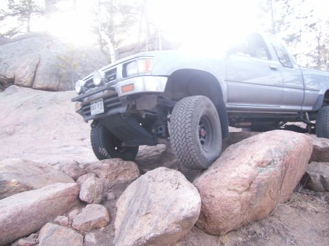 1993 Ford Bronco - A silver 1993 Ford Bronco is shown navigating a rocky terrain, with its front tire placed on a large boulder. The vehicle's front end, incl…