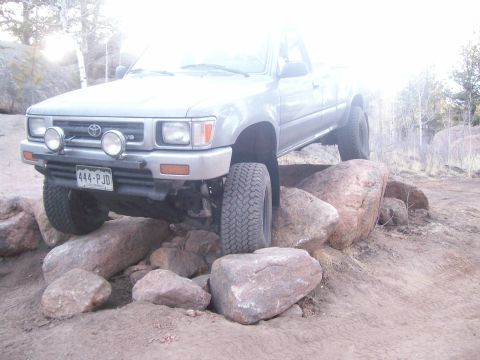 1993 Ford Bronco - A silver Toyota pickup truck is positioned on large rocks, with its front tire elevated. The truck features two large fog lights mounted on…
