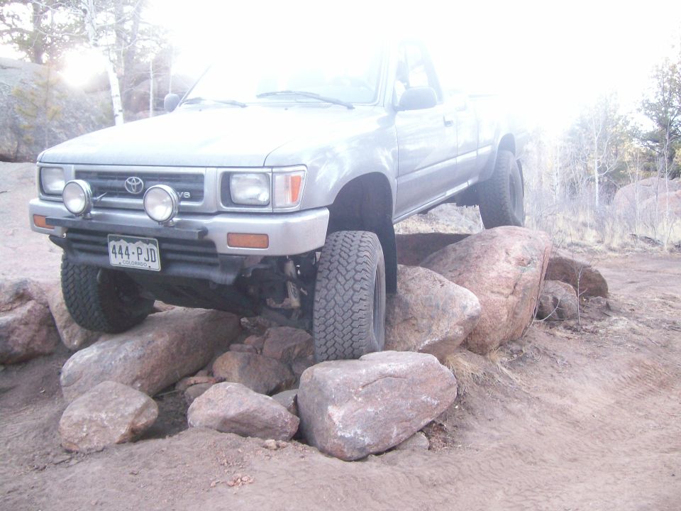 Silver Toyota pickup truck on rocks, front tire elevated, with fog lights and license plate.
