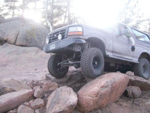 1993 Ford Bronco - A brown 1993 Ford Bronco is shown off-roading over large rocks, with its front tire elevated. The sun is shining brightly through the trees…