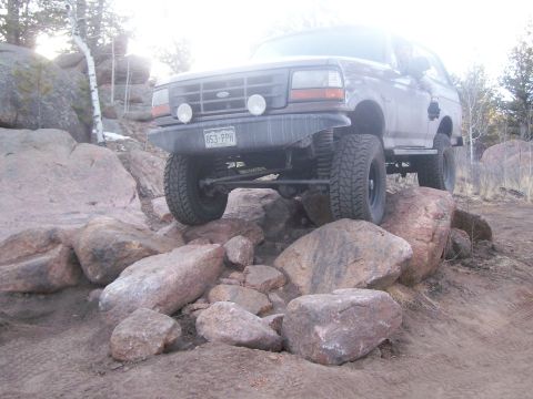 1993 Ford Bronco - A brown 1993 Ford Bronco is shown off-roading over large rocks on a dirt trail. The front of the vehicle is angled towards the camera, with…