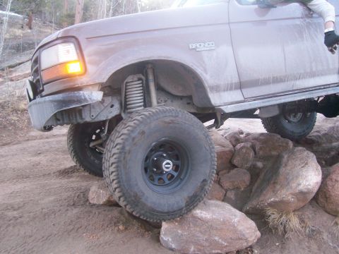 1993 Ford Bronco - The front fender and wheel of a brown Ford Bronco are shown navigating over large rocks on a dirt trail. The vehicle's orange turn signal i…