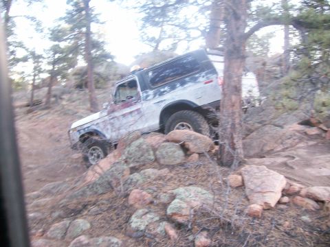 1993 Ford Bronco - A white and blue Ford Bronco is navigating a rocky, wooded trail. The vehicle is angled upwards on the trail, with its front tire on a rock.