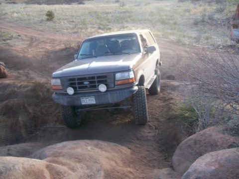 1993 Ford Bronco - A brown 1993 Ford Bronco is driving off-road on a dirt path, angled towards the viewer. The vehicle is navigating a steep incline with larg…