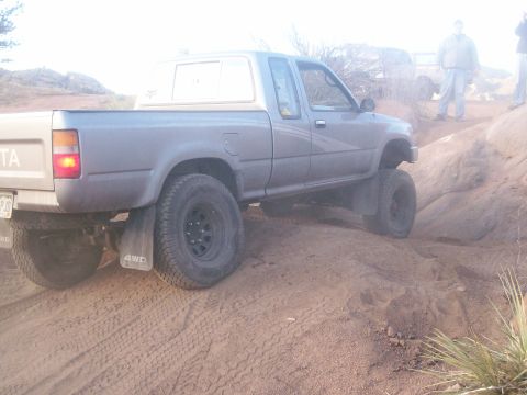 1993 Ford Bronco - The rear passenger side of a lifted, gray Toyota pickup truck is shown driving on a dirt trail. The truck has large off-road tires and "4WD…