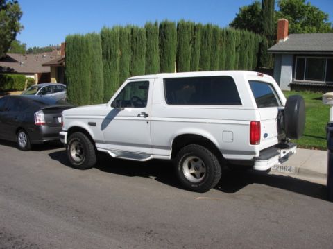 1988 Ford Bronco - The side view of a white two-door Ford Bronco is parked on a residential street. A dark-colored hybrid car is parked in front of it, and a…