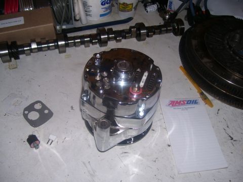 1990 Ford Mustang - A chrome alternator sits on a workbench next to a camshaft, a clutch assembly, and a notepad with an Amsoil logo. Several small electrical…
