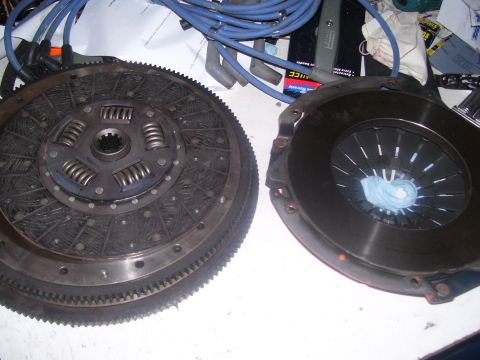 1990 Ford Mustang - A clutch disc and pressure plate are laid out on a white surface, likely for a 1990 Ford Mustang. Blue spark plug wires are visible in the…