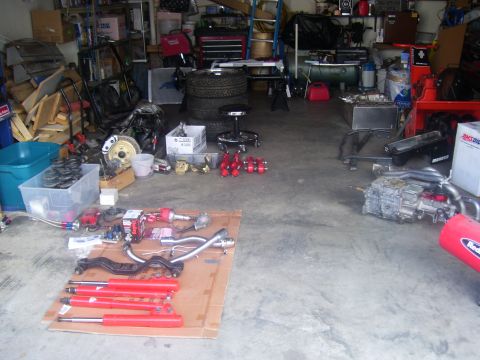 1990 Ford Mustang - A collection of automotive parts, including a transmission, exhaust components, and suspension parts, are laid out on a concrete garage flo…