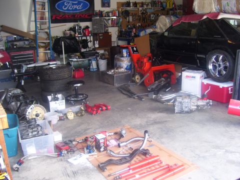1990 Ford Mustang - A black 1990 Ford Mustang is parked in a cluttered garage next to a snowblower and various car parts. The car has chrome wheels and appears…