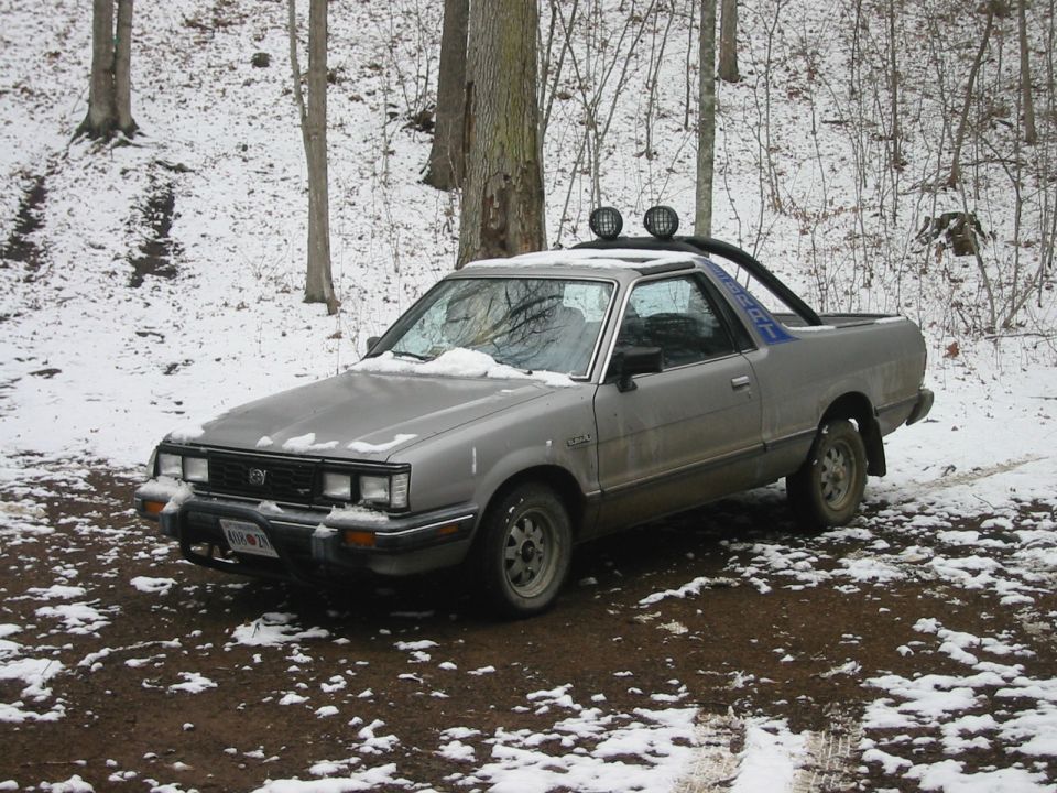 Silver Subaru Brat with roll bar and lights in a snowy, wooded area.
