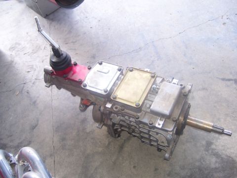 1990 Ford Mustang - A manual transmission is shown mounted on a concrete floor. The transmission features a red housing near the shifter, with multiple metal a…