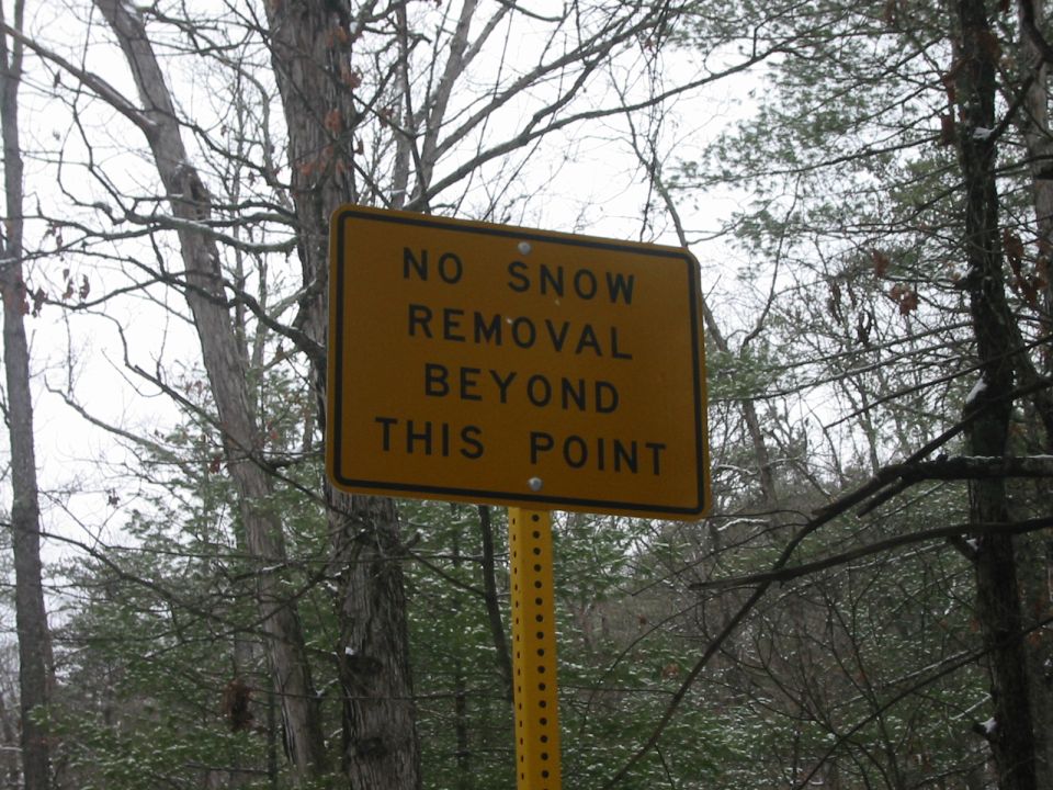 Sign that reads "NO SNOW REMOVAL BEYOND THIS POINT" in a wooded area.