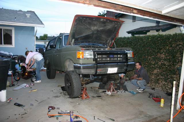 Green Ford Bronco with hood open, being worked on by two people. Tools on driveway.