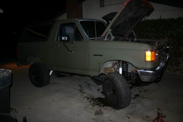 Lifted olive green 1991 Ford Bronco with hood open, parked at night.