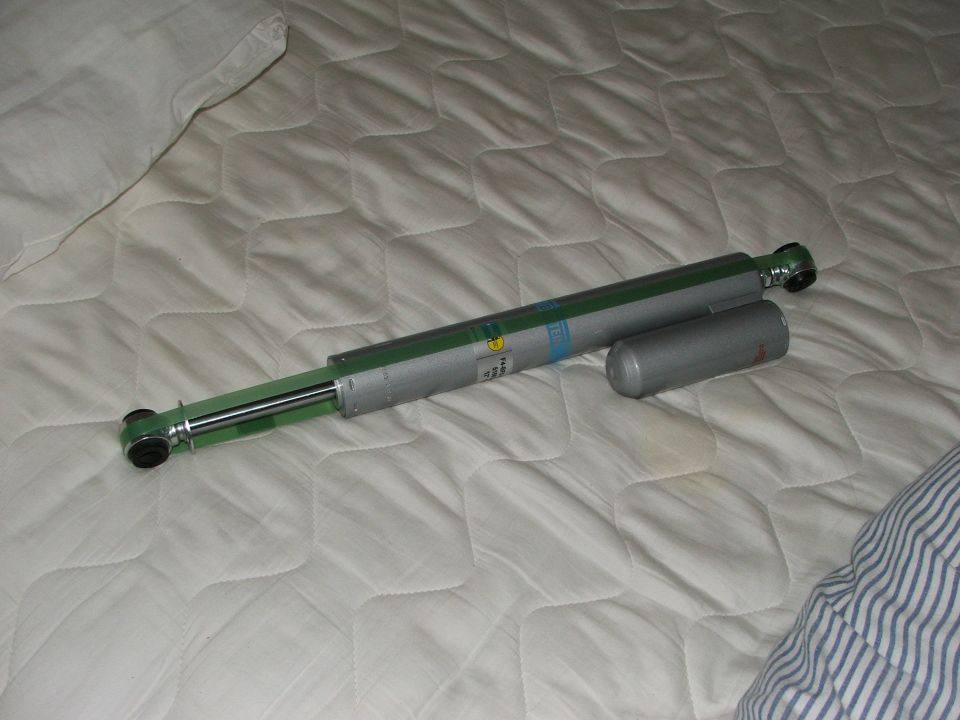 Silver and green Bilstein shock absorber with separate reservoir on a white quilted surface.