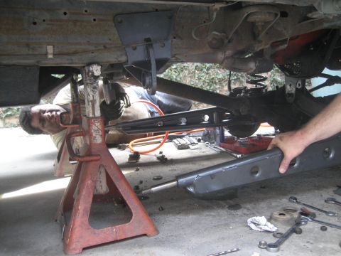 1987 Ford Bronco - A mechanic works on the undercarriage of a vehicle supported by red jack stands, with a differential and suspension components visible.