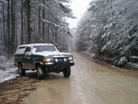 1989 Ford Bronco - A green Ford Bronco with a white removable top sits at the edge of a snow-dusted dirt road in a pine forest during falling snow, with snow…