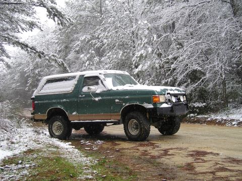 1989 Ford Bronco - A green Ford Bronco is parked on a dirt road in a snowy forest. The vehicle is covered in a light dusting of snow, and the trees surroundin…