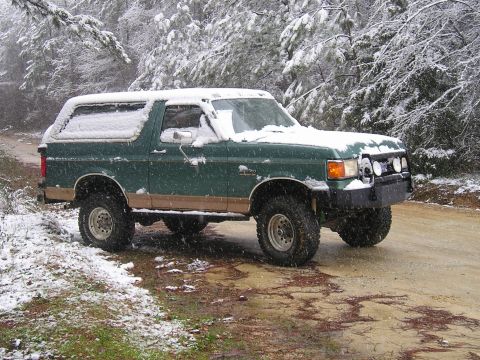 1989 Ford Bronco - A green Ford Bronco covered in snow sits on a dirt road surrounded by snow-covered trees. The vehicle has a tan lower body trim and appears…