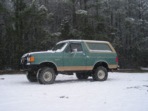 1989 Ford Bronco - Driver's-side profile of a lifted green Ford Bronco with a tan hardtop and tan lower two-tone parked in falling snow, snow settled along th…