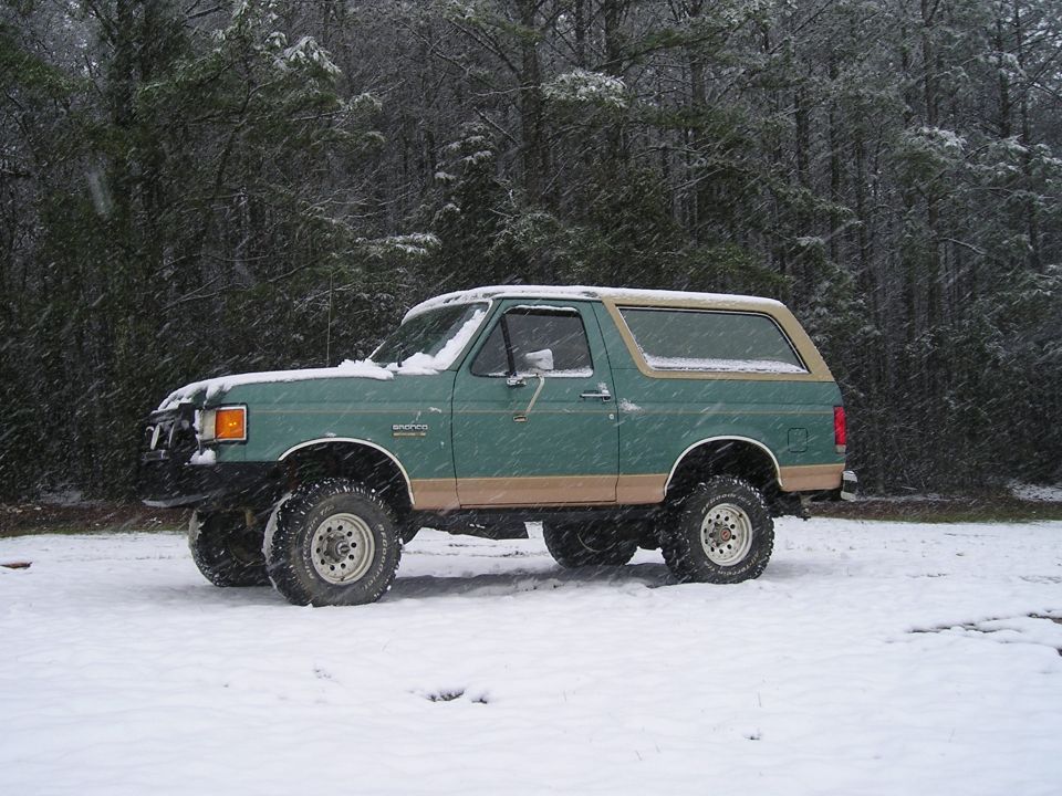 Green and tan Ford Bronco parked in falling snow, driver's side profile