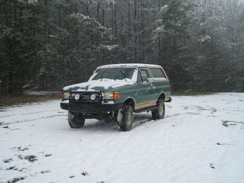 1989 Ford Bronco - A green Ford Bronco sits in a snowy clearing with a forest in the background. Snow is falling heavily.