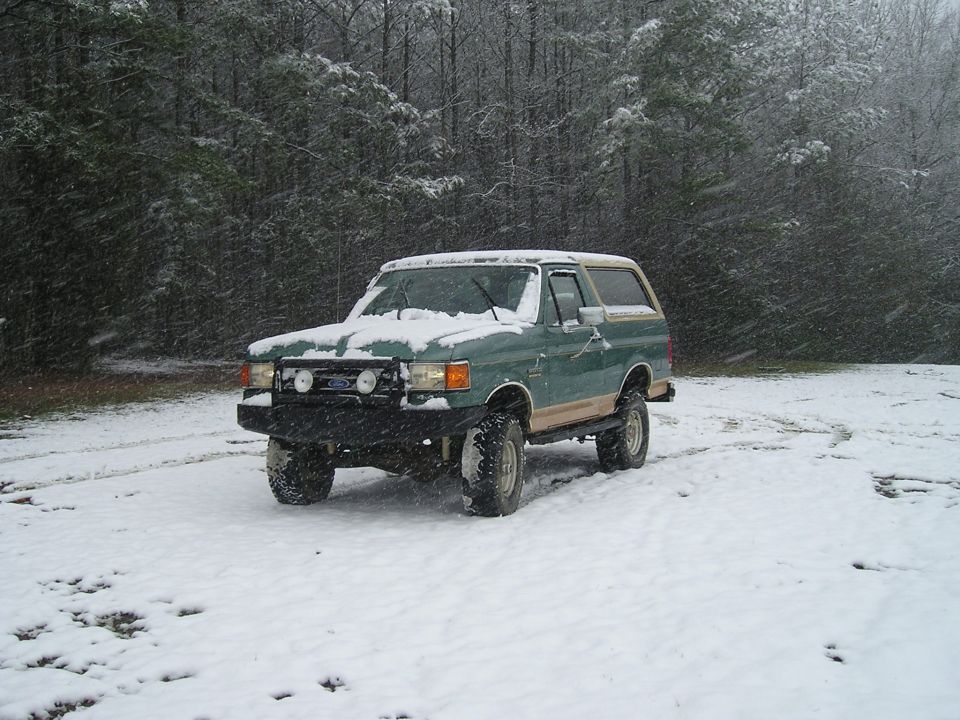 Green Ford Bronco in a snowy forest clearing during a snowstorm.