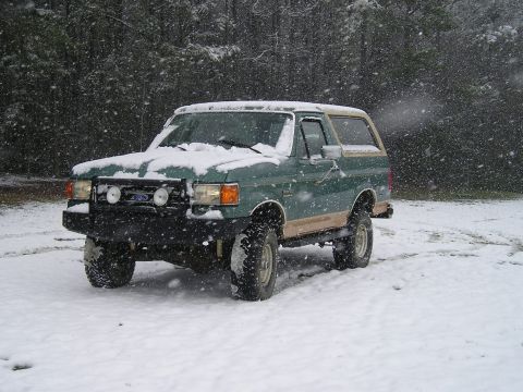 1989 Ford Bronco - A green and tan Ford Bronco sits parked in a snowy landscape with snow falling. The front of the vehicle features a black bull bar with two…